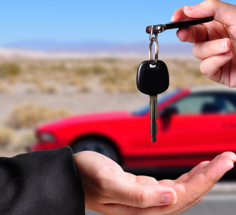 Image of a car key being given to another person, hands only visible with a red car in the background